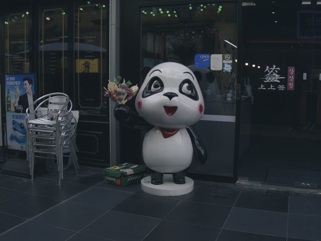 인천 송도 트리플스트리트의 한 가게 앞에 설치된 판다 조형물. 하얀 몸에 검은 팔과 다리, 빨간 볼이 돋보이는 귀여운 캐릭터가 꼬치다발을 들고 환한 표정을 짓고 있다. 배경에는 유리문과 한글·영문 간판이 보이며, 옆에는 금속 의자가 쌓여 있고 벽면에는 포스터가 부착되어 있어 상업 공간의 활기찬 분위기를 더한다.