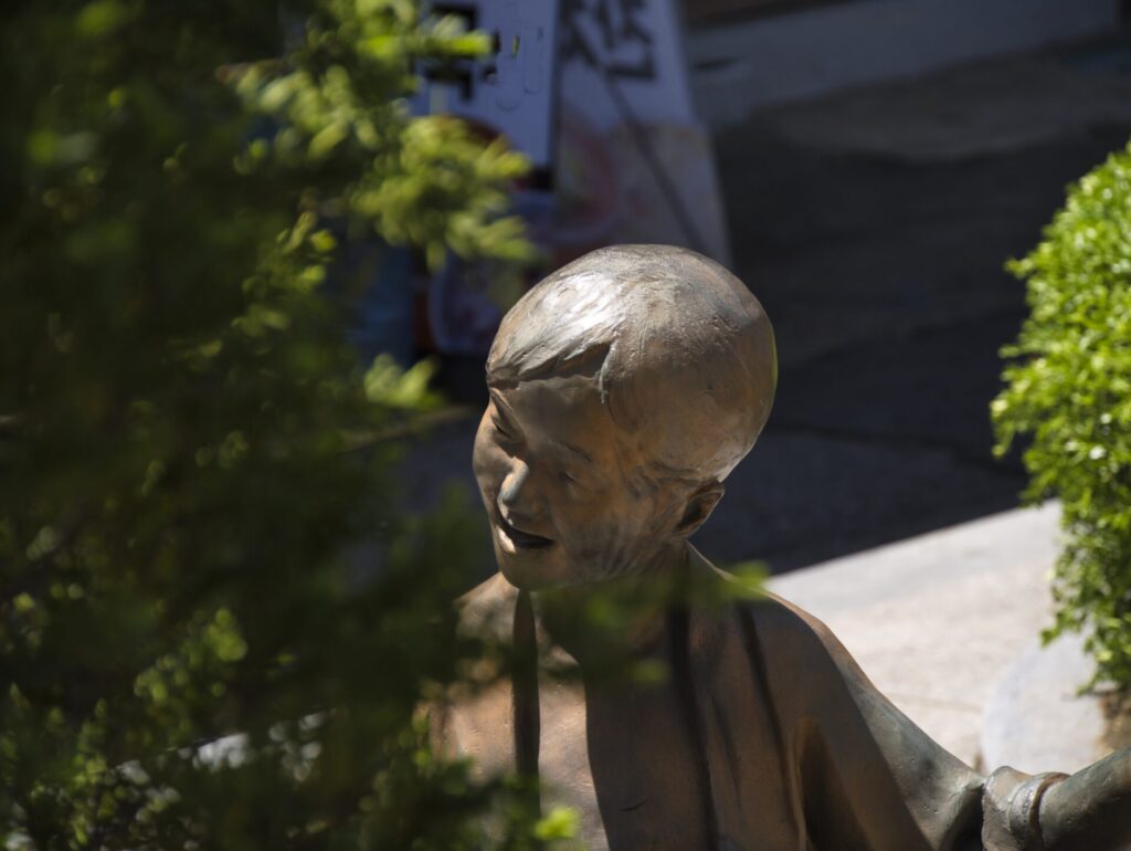인천 송현시장 한켠에 자리한 아이 동상. 해맑은 표정을 지닌 청동 조형물이 푸른 식물 사이에 살짝 가려져 있으며, 햇살 아래 반짝이는 질감이 생동감을 더한다. 배경에는 시장 간판과 도시적 요소들이 어우러져 지역의 정서와 일상의 풍경을 함께 담아낸다.