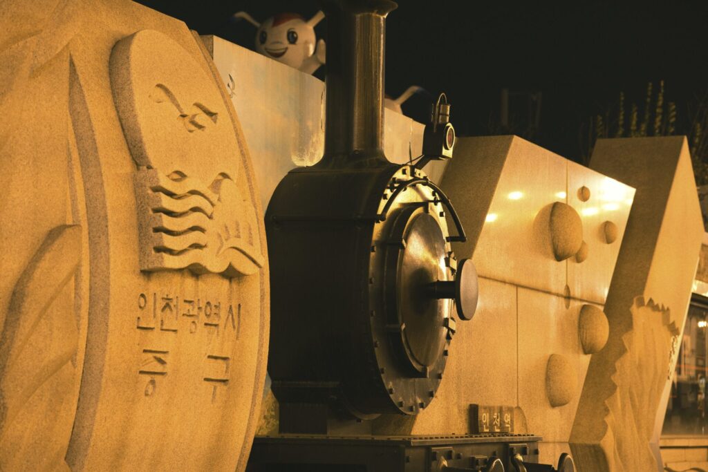 인천 중구청 앞에 설치된 기차 모형 조형물. 증기기관차를 형상화한 석재 구조물로, 원형 보일러와 굴뚝, 볼트 장식이 사실적으로 표현되어 있다. 측면에는 파도와 갈매기, 돛단배가 새겨진 문양과 ‘인천광역시 중구’라는 글씨가 함께 조각되어 지역의 상징성을 담고 있다. 야간 조명 아래 따뜻한 빛이 조형물의 질감을 강조하며, 도시의 문화적 풍경을 만들어낸다.