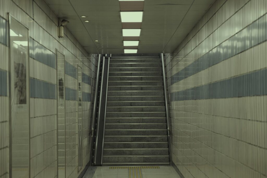 인천 도원역 지하도에서 촬영한 내부 풍경. 베이지색 타일 벽면과 파란색 띠 장식이 깔끔하게 정돈되어 있으며, 중앙 계단은 금속 손잡이와 시각장애인을 위한 점자 블록이 설치되어 있다. 천장에는 직사각형 형광등이 일렬로 배치되어 밝은 조명을 제공하고, 왼쪽 벽면에는 CCTV가 설치되어 안전한 분위기를 조성한다. 대칭적인 구조가 도시 지하 공간의 질서와 기능미를 잘 보여준다.