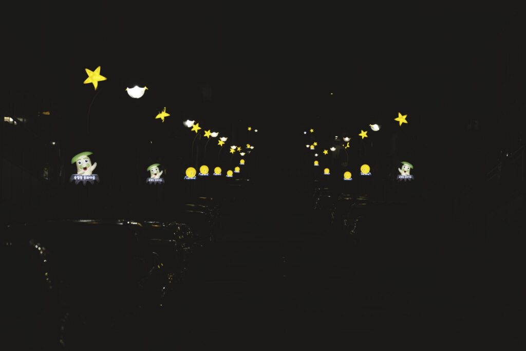 야간에 촬영한 인천 동화마을의 가로등 풍경. 노란 별 모양과 캐릭터 얼굴이 장식된 가로등들이 거리 양쪽에 줄지어 서 있으며, 어두운 배경 속에서 따뜻한 빛을 발하며 동화 같은 분위기를 자아낸다. 가로등 불빛이 주차된 차량에 반사되어 도시 속의 마법 같은 순간을 연출한다.