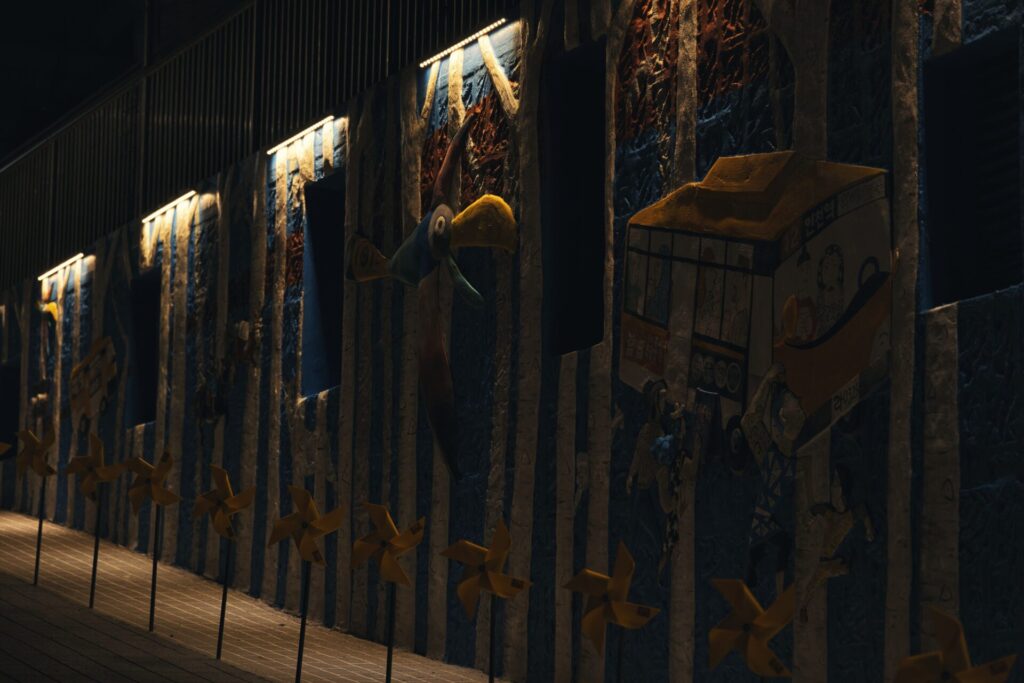 야간에 촬영한 인천 동화마을의 벽화. 파란색과 흰색 줄무늬 벽면 위에 커다란 부리의 새, 만화 스타일의 버스, 다양한 캐릭터들이 입체적으로 표현되어 있으며, 조명이 일부 요소를 강조해 환상적인 분위기를 연출한다. 앞쪽에는 노란 바람개비들이 줄지어 서 있어 동화 같은 장면에 생동감을 더한다.