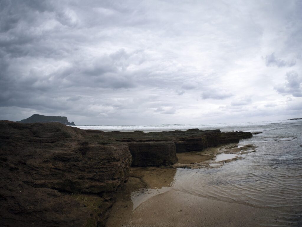 제주도 광치기 해변에서 촬영한 흐린 날의 해안 풍경. 울퉁불퉁한 암석 지형이 앞쪽에 펼쳐지고, 얕은 바닷물이 그 사이를 채우며 자연의 질감을 강조한다. 회색빛 구름이 하늘을 뒤덮고, 멀리 바다로 뻗은 육지가 수평선을 따라 시야를 이끈다. 거친 바위와 흐린 하늘이 어우러져 제주도의 고요하면서도 강렬한 자연미를 담아낸다.