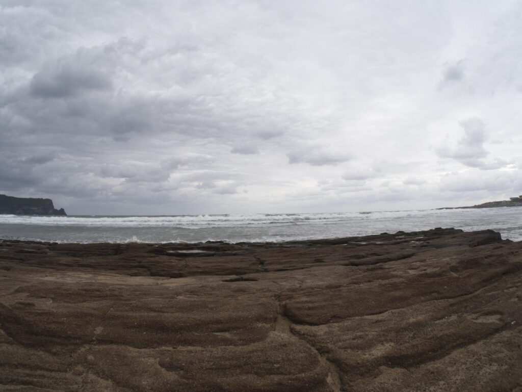 제주도 광치기 해변에서 촬영한 풍경. 앞쪽에는 층층이 쌓인 바위 지형이 펼쳐지고, 멀리에는 짙은 구름 아래 잔잔한 바다와 수평선이 이어진다. 왼쪽에는 어두운 절벽이 바다로 뻗어 있으며, 흐린 하늘과 대비되는 해안선이 제주도의 거친 자연미를 강조한다.