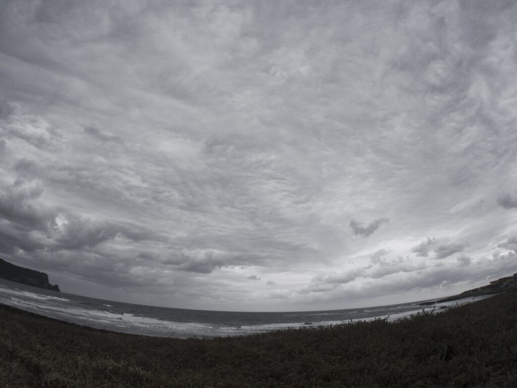 제주도 광치기 해변에서 촬영한 흐린 하늘과 해안 풍경. 두터운 회색 구름이 하늘을 가득 메우고, 잔잔한 파도가 해변을 따라 밀려온다. 왼쪽에는 어두운 바위 절벽이 바다로 뻗어 있으며, 앞쪽에는 풀과 관목이 해변을 향해 펼쳐져 있다. 광활한 하늘과 자연의 질감이 어우러져 제주도의 거친 아름다움을 담아낸다.