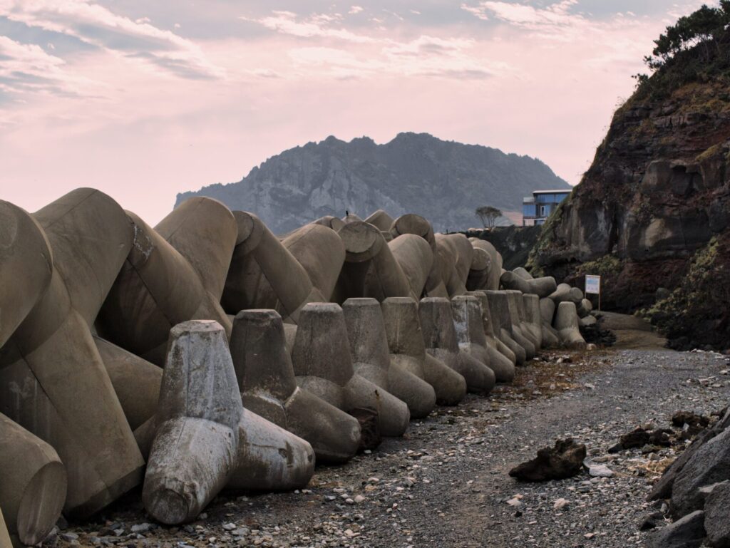 제주도 바닷가에서 촬영한 해안 풍경. 해안선을 따라 대형 콘크리트 테트라포드가 줄지어 배치되어 있으며, 파도의 힘을 흡수해 해안을 보호하는 역할을 한다. 배경에는 바위 절벽과 푸른 식생, 그리고 절벽 끝에 자리한 작은 파란 건물이 보인다. 멀리 산맥이 펼쳐지고, 분홍빛이 감도는 하늘이 일출 또는 일몰의 분위기를 더하며, 자연과 인공 구조물이 조화를 이루는 인상적인 장면을 연출한다.