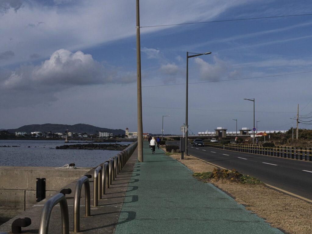 제주도 해변가에서 촬영한 해안 도로 풍경. 왼쪽에는 바다와 콘크리트 방파제가 이어지고, 금속 난간 너머로 잔잔한 물결이 보인다. 초록색 보행로에는 사람이 걷고 있으며, 오른쪽 도로에는 가로등과 차량이 멀리 보인다. 배경에는 건물과 언덕이 펼쳐지고, 하늘에는 얇은 구름이 흩어져 제주도의 여유로운 분위기를 자아낸다.