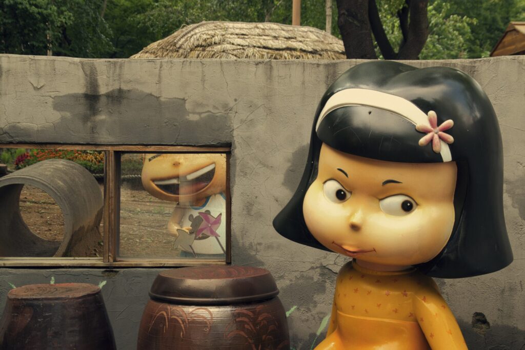 인천 솔빛 공원에서 촬영한 귀여운 인형상. 앞쪽에는 장난기 어린 표정을 짓고 있는 검은 머리 소녀 인형이 노란색 물방울 무늬 드레스를 입고 서 있으며, 뒤쪽 콘크리트 벽 너머로는 보라색 불가사리를 든 웃고 있는 아이 인형이 보인다. 주변에는 커다란 항아리와 자연 소재의 구조물이 어우러져 독특하고 유쾌한 분위기를 자아낸다.