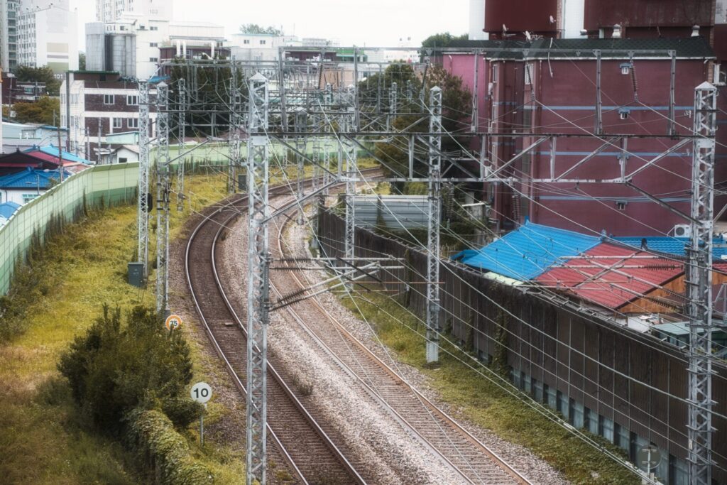 인천역 인근의 전철길을 따라 굽이치는 철로 풍경. 왼쪽에는 녹색 펜스와 식생이 이어지고, 오른쪽에는 붉은 건물과 파란 지붕이 어우러진 도시 구조물이 자리잡고 있다. 전철 전선과 전봇대들이 철로를 따라 설치되어 있는 등 도시 철도 시설의 세부적인 모습이 돋보인다.