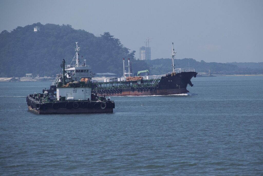 인천 월미도 앞바다에서 촬영한 선박 풍경. 앞쪽에는 튼튼한 구조의 예인선이 항해 중이며, 뒤쪽에는 대형 화물선이 잔잔한 수면 위에 떠 있다. 배경에는 숲으로 둘러싸인 해안과 건물들이 보이며, 흐릿한 하늘 아래 평화롭고 산업적인 분위기가 어우러진다.