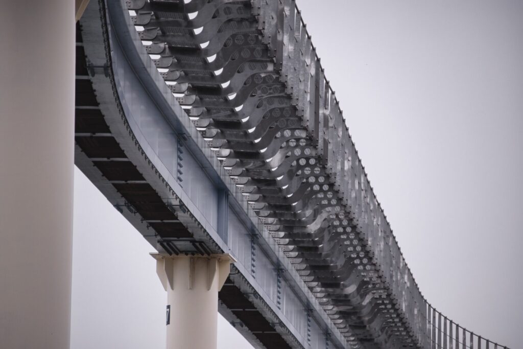 인천 월미도 모노레일의 구조물을 아래에서 올려다본 모습. 굵은 원형 기둥들이 모노레일을 지탱하고 있으며, 곡선 형태의 금속 지지 구조가 반복적으로 배열되어 독특한 패턴과 대칭미를 이룬다. 흐린 하늘을 배경으로 현대적인 도시 인프라의 세련된 디자인이 돋보인다.