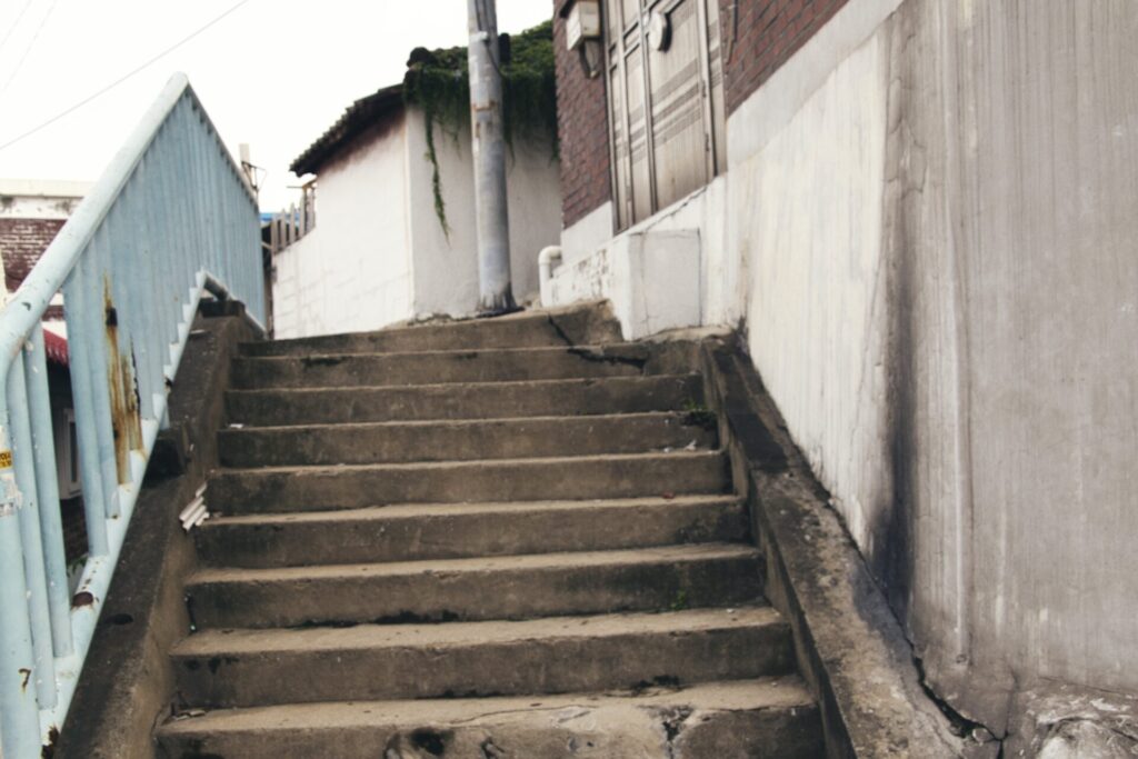 재개발을 앞둔 인천 구도심의 낡은 콘크리트 계단. 푸른 철제 난간과 얼룩진 벽면이 세월의 흔적을 보여주며, 계단 위로는 벽돌 건물과 전봇대가 보인다. 주변의 오래된 주택들과 함께 도시의 과거와 변화의 경계에 선 풍경을 담고 있다.